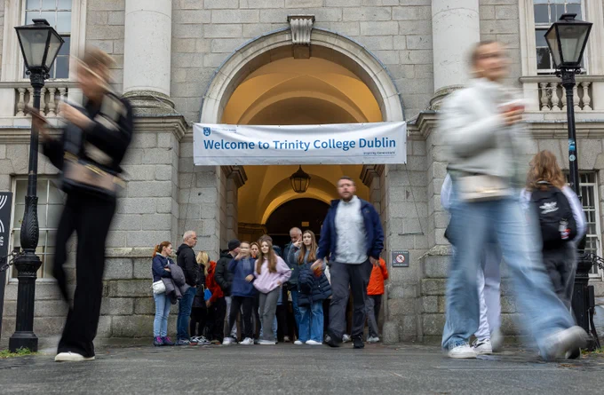 Blurred figures walking by the entrance of Trinity College Dublin with a 'Welcome to Trinity College Dublin' banner above the archway.