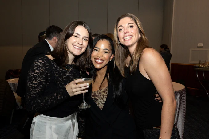 Three women in formal attire stand together smiling at the camera