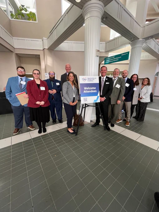Nate Cox, Brynne Olsen, Matthew Tekaucic, Michael Hoag, Kristen Ballinger, OOD Director Kevin Miller, Brian Crowe, Chris Brown, Jennifer Finnerty, and Nikki Hall. A sign in the center of the group reads "Welcome attendees."