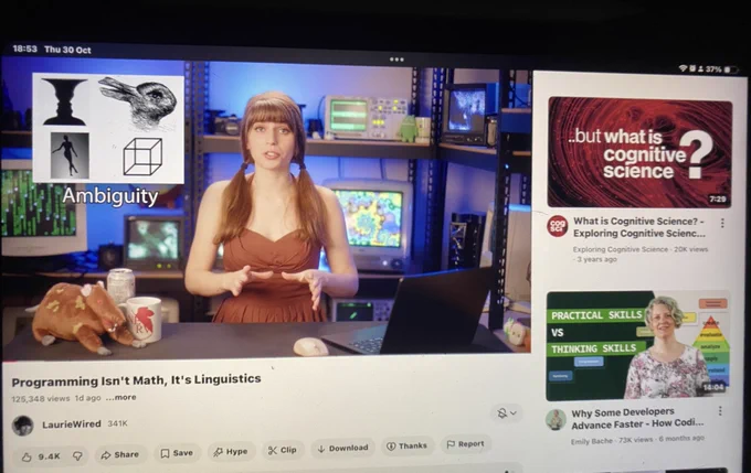 Tela do YouTube aberta em um iPad. À esquerda, um vídeo com uma apresentadora em um estúdio tecnológico, com ícones de fala. Na mesa há um boneco de pelúcia marrom, caneca e objetos pequenos. Abaixo do vídeo, o título “Programming Isn’t Math, It’s Linguistics” e botões de interação. À direita, coluna de vídeos recomendados com miniaturas e textos como “What is Cognitive Science?” e “Practical skills vs thinking skills”. Barra superior mostra horário “18:47 Thu 30 Oct”.