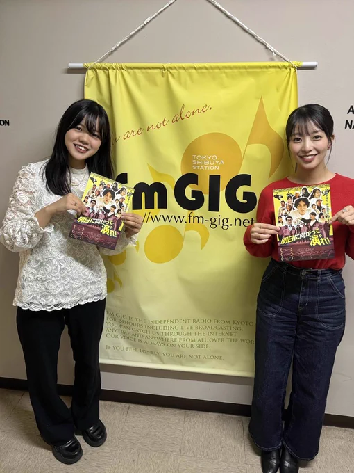 「fm GIG」と書かれた黄色いタペストリーを挟んで右に野口、左に熊倉が立っている。
「あすに向かってやれ」のフライヤーを手に持っている。