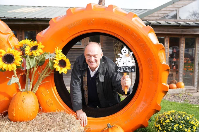 Ed Davey smiling through a pumpkin-themed tractor tyre, holding a ‘Happy Halloween’ sign.