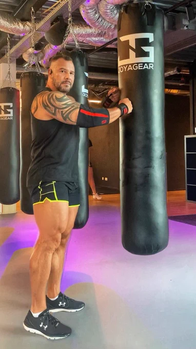 Man in a sleeveless black shirt and short neon-trim spandex shorts standing by a punching bag in the gym, muscles tensed and focused.