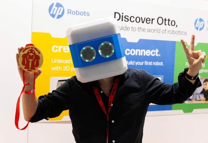 A person wearing a robot-shaped helmet poses with a peace sign and holds a "Maker of Merit 2025" medal at the HP Robots booth during Maker Faire Rome. Behind them is a colorful display promoting Otto, a robotics education project.