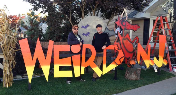 Mark Sceurman and Mark Moran stand on a lawn in front of a suburban home decorated for Halloween. They are standing behind a giant sign that looks like the Weird NJ magazine logo. 