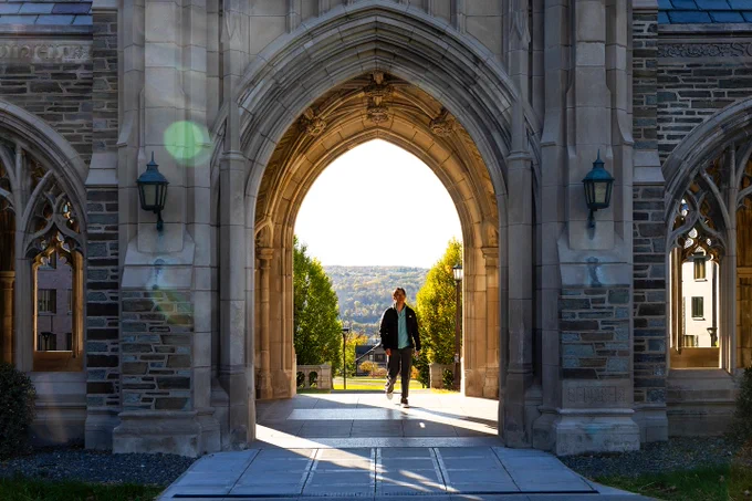 A person walks through the sunlit archway of the Cornell War Memorial entrance with a view of the distant landscape visible in the background.