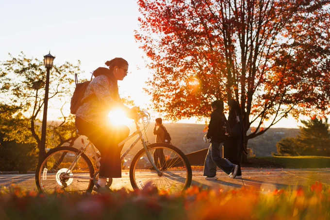 Person on a bike pauses on a Cornell University path during a sunset, while other people walk by, with vibrant autumn trees in the background.