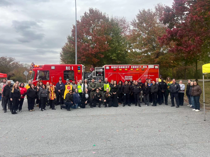 Group photo of Montgomery County emergency responders and other community members standing in front of a red fire engine with autumn trees in the background at the Public Safety Trunk or Treat event.