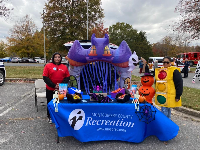 Montgomery County Recreation's vehicle trunk decorated for Halloween with an inflatable bat and ghost and a table filled with candy and two staff members standing alongside it.