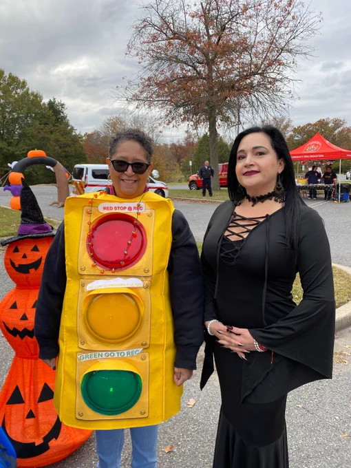 A Montgomery County Recreation staff member dressed in a colorful traffic light costume standing next to another person in costume in a long black dress with lace-up detail at the Public Safety Trunk or Treat event.