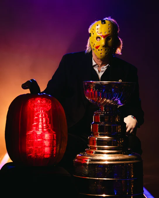 Photo of the Stanley Cup next to a large pumpkin with the Stanley Cup's likeness carved on it. Behind them, the Keeper of the Cup Phil Pritchard sits with a mask on.