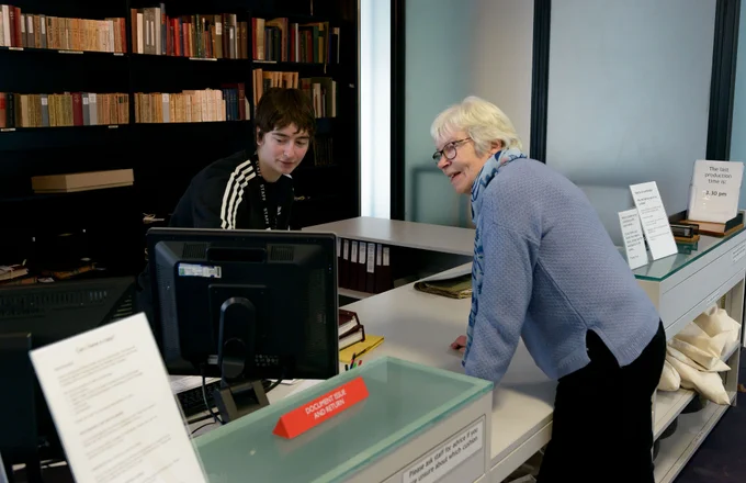 From behind a reception desk, a young person wearing a staff lanyard shows an older person something on a computer monitor. Behind them stands a large shelving unit filled with books.