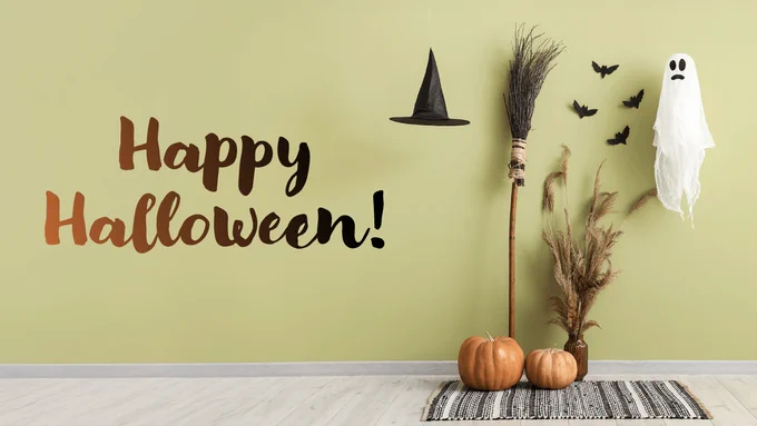 Halloween-themed decoration with 'Happy Halloween' text on the wall, two pumpkins on the floor, a broomstick, dried grass, a hanging ghost figure, a witch hat, and flying bat cutouts.