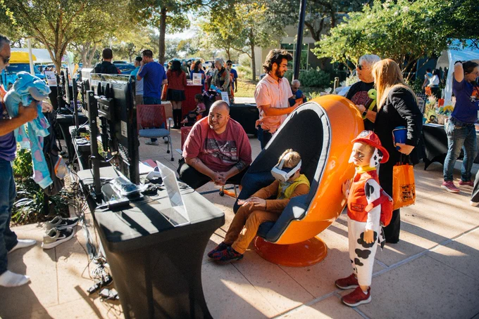 People enjoying a community event with various entertainment setups, including video games and unique seating arrangements, on a sunny day. Some attendees are dressed in casual outfits and others in costumes, enhancing the festive atmosphere.