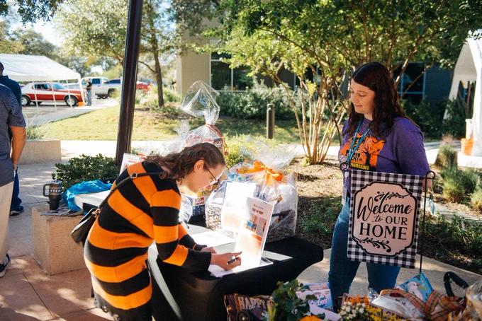 Person in a costume signing up at an outdoor event with various gift baskets on the table, overseen by another person, near a 'Welcome to Our Home' sign.