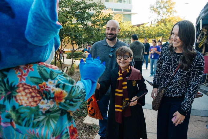 Person in a vibrant floral Stitch costume greeting a family of three, including an excited child dressed in a Hogwarts scarf, in a sunny outdoor setting.