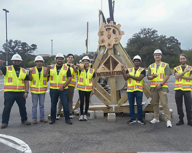 A group of students wearing safety helmets and reflective vests standing in front of a wooden trebuchet with an International Club logo.