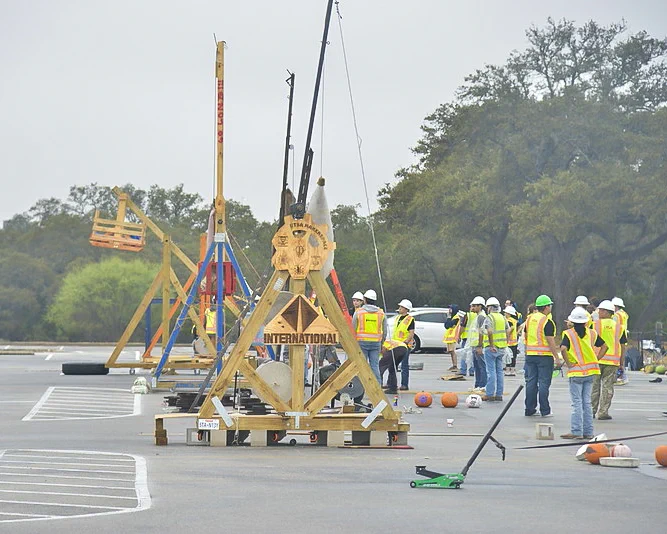 Students in safety vests and helmets are assembling large trebuchets in the parking lot, surrounded by pumpkins.