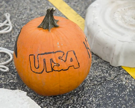 Orange pumpkin with "UTSA" carved into it, placed on a textured ground of a parking lot.