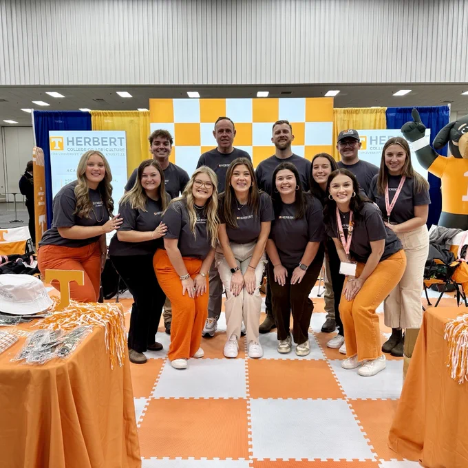 Group of people smiling in front of a University of Tennessee promotional booth decorated in orange and white with the school's mascot visible.