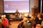 A woman speaking on stage in front of an audience with a screen behind her displaying 'FALL INTO PLACE'.