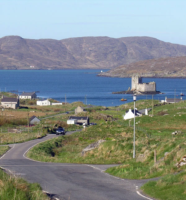 Today's 'Where in Scotland' image. It is a squarish image showing a view down a narrow single track road that enters the bottom centre of the frame before heading downhill to the left then back to the right into a scattered settlement of houses and cottages on the shore of the sea, which is blue under blue skies. In the middle right of the frame is a castle on its own islet, while in the distance, more land rises to rough hills.