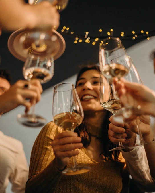 Person smiling and toasting with a glass of wine, surrounded by others, in a festive atmosphere with blurred lights in the background.
