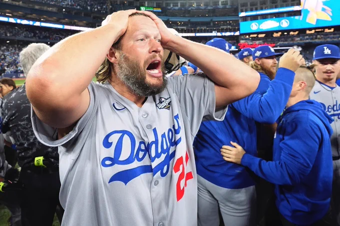 Clayton Kershaw holds his hands on his head in amazement after winning the World Series. He wears a road gray Dodgers uniforms with blue and red lettering.