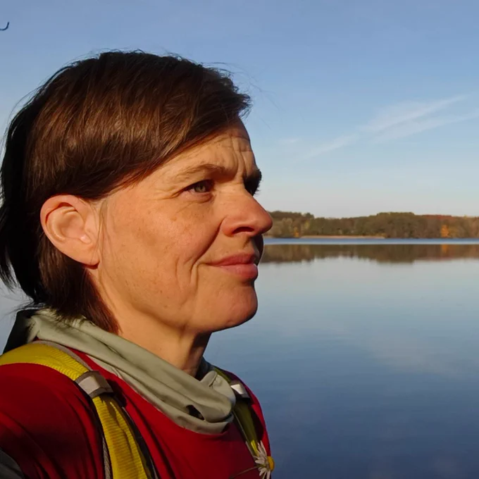Selbstportrait der lächelndes Komplizin vor einem See. Ihr morgensonnenbeschienenes Gesicht im Halbprofil ist in der linken Bildhälfte zu sehen. Sie blickt beglückt und versonnen über den See, in dessen glatter Wasseroberfläche blauer Himmel mit einer weißen Federwolkenschliere sowie das herbstlaubbunt waldgesäumte Ufer im Hintergrund sich spiegeln.  Sie trägt ein rotes Shirt und ein salbeifarbiges Halstuch, über Brust und Schultern laufen gelbe Rucksackgurte. Das mittelblonde glatte Haar ist halblang und hinter das rechte Ohr gestrichen. 
