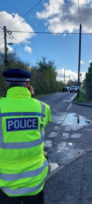 This photo shows a police officer wearing a high visibility jacket holding a speed gun, aiming it down a long stretch of country road.