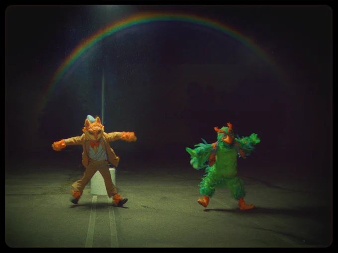 A pair of dancers portraying anthropoorpic creatures caper through a parking lot at night under a rainbow