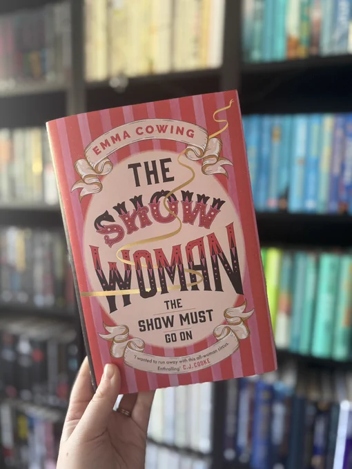 Hand holds "The Show Woman" book by Emma Cowing with red striped cover. Blurred bookshelf background.