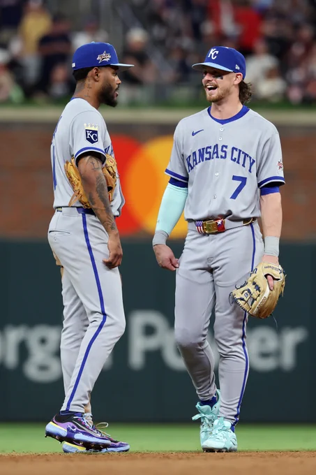 A photo of Maikel Garcia and Bobby Witt Jr. standing and talking in the infield. They both wear road gray Kansas City Royals uniforms with blue KC caps.