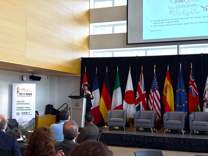 IMAGE: Photo prise pendant la conférence internationale R7 .  Une personne au podium sur la scène présente devant une foule. On voit derrière plusieurs drapeaux et un écran géant.