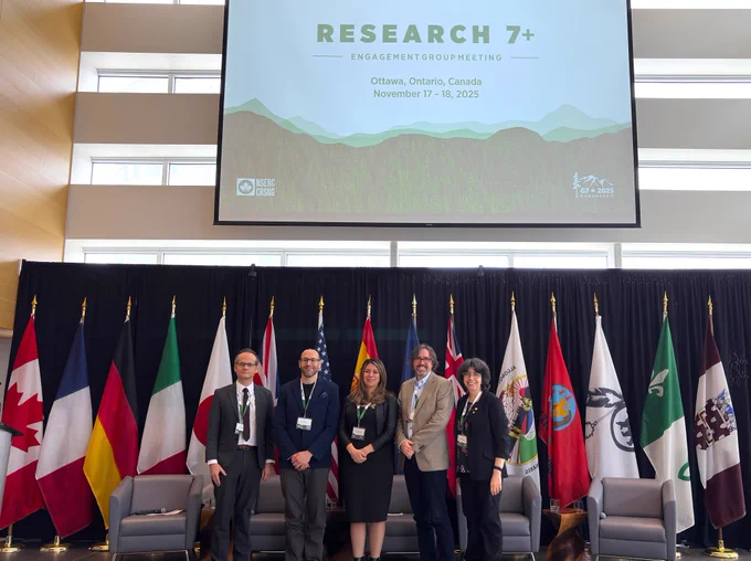 IMAGE: Photo prise pendant la conférence internationale R7 .  Cinq personnes posent pour un portrait officiel sur la scène. On voit derrière plusieurs drapeaux, des fauteuils et un écran géant sur lequel on peut lire "RESEARCH 7 ".