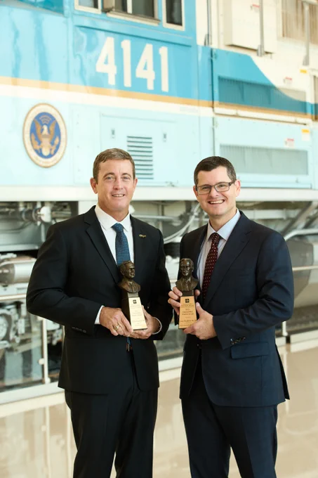 LtCol (Ret) Travis Patterson ’98 in suit and blue tie holding outstanding alumni award next to Casey Blackburn ’99, ’03 in suit with red tie and eye glasses holding outstanding alumni award standing infront of bush 41 train 
