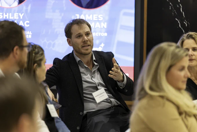 Man in business attire, sitting as part of an audience, speaks to someone off camera