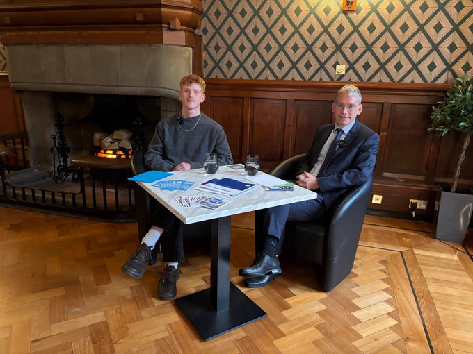 Two people sit across from each other at a small table in an elegant wood-paneled room with a lit fireplace in the background. The person on the left is SRC president Duncan Calvert. The person on the right is the University of Glasgow's new Principal and Vice-Chancellor, Professor Andy Schofield. Papers, brochures, and two glasses of water are on the table between them. 