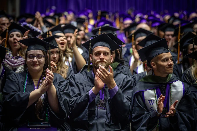 A female graduate and two male graduates clap as they sit in the front row at a recent Weber State commencement ceremony.