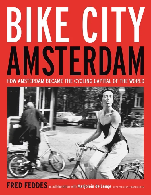 Red book cover.  Top half, all in capitals, white "BIKE CITY", black "AMSTERDAM", small white "HOW AMSTERDAM BECAME THE CYCLING CAPITAL OF THE WORLD".  Bottom half, black and white photo of a woman cycling, sticking her tongue out towards the photographer, above black "FRED FEDDES" and white "in collaboration with Marjolein de Lange - UITGEVERIJ BAS LUBBERHUIZEN".
