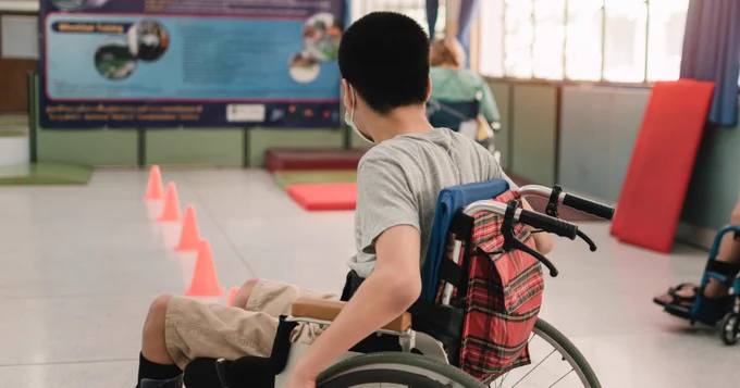 A young boy with short black hair facing away from the camera. He is using a manual wheelchair and is facing towards a line of pylons set up on the floor
