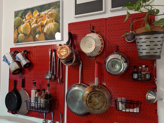 A red Wall Control metal pegboard mounted on a kitchen wall, neatly holding pans, strainers, utensils, spices, moka pots, and a hanging plant, showcasing an organized and efficient cooking setup.