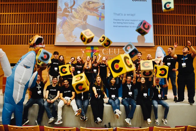 Foto de grupo del equipo organizador y voluntariado en el auditorio, lanzando al aire varias cajas de colores con el logo de Commit, celebrando el final del evento.