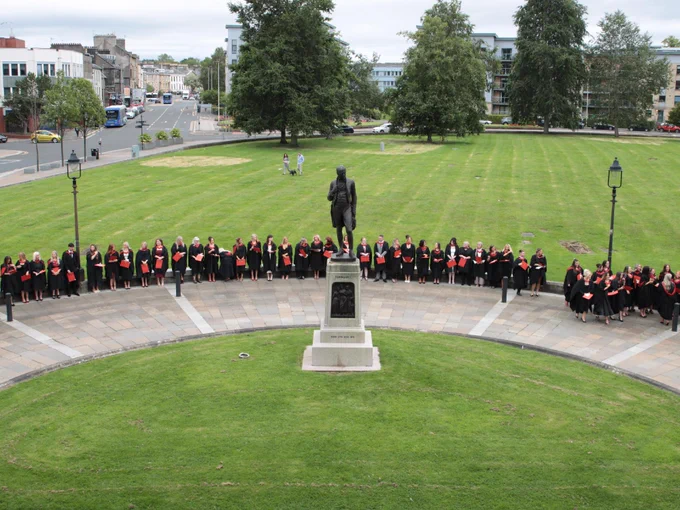 Graduates in traditional black and red academic regalia gathered in a semi-circle around a statue on, with green lawns and city buildings in the background.