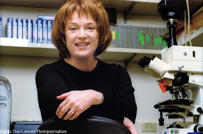 Linda Buck sitting in a laboratory with a microscope and files in the background, smiling at the camera.