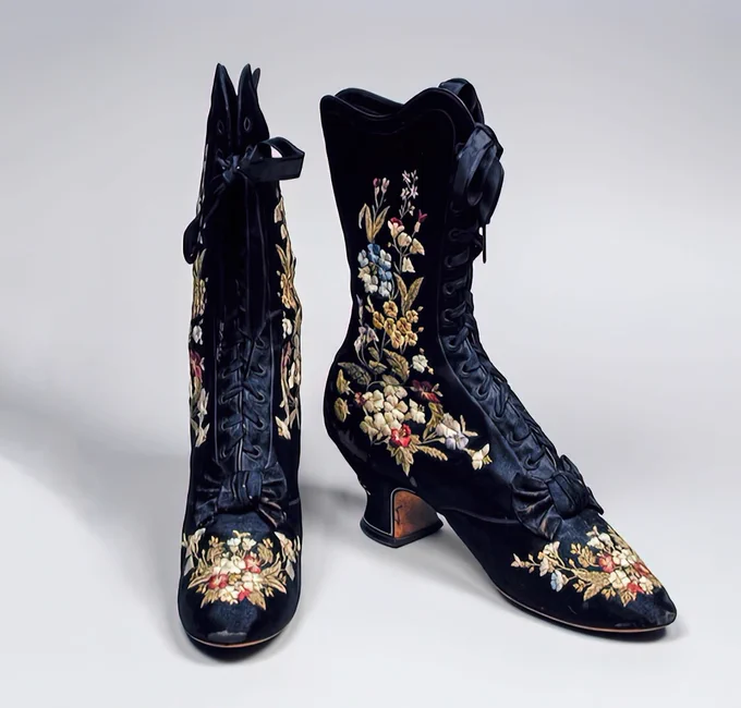 Photograph of a pair of dark midnight blue silk ankle boots with a rounded pointed toe and Louis heels. The boots lace up to the top and the silk is covered with multi coloured floral embroidery
