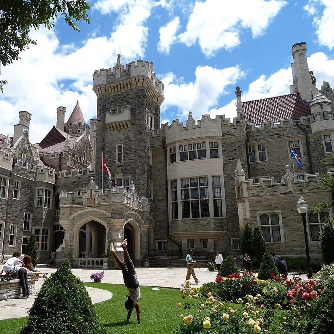 A grand stone castle with multiple towers, turrets, chimneys, and arched windows stands under a partly cloudy blue sky. Canadian flags fly on the building. In the foreground, a person performs a handstand on green grass near a paved path, while others walk or sit nearby. Well-maintained gardens feature colorful roses in red, pink, and yellow, conical evergreen shrubs, a small fountain with an orange traffic cone, and trees framing the scene.