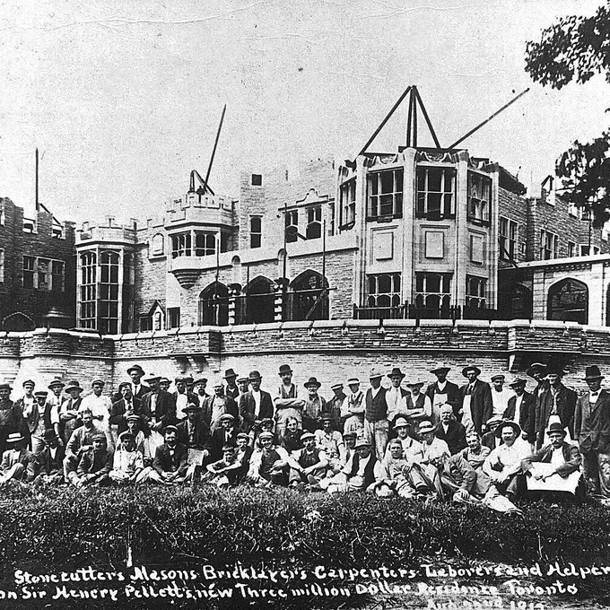 Black-and-white historical photograph of a large group of men—stonecutters, masons, bricklayers, carpenters, laborers, and helpers—posed in rows, seated and standing on grass in front of a partially constructed grand stone mansion with towers, arched windows, and scaffolding. The men wear early 20th-century work clothes and hats. Handwritten text at the bottom reads: "Stonecutters Masons Bricklayers Carpenters Laborers and Helpers on Sir Henry Pellatt's new Three million Dollar Residence Toronto."