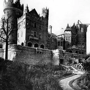 Black-and-white historical photograph of a large Gothic-style castle with tall round tower, battlements, multiple chimneys, arched windows, and stone walls. A curved pathway winds up a grassy hill toward the building, flanked by bare trees and overgrown vegetation. The image has a vintage quality with high contrast and visible grain.