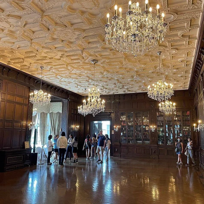 Interior photo of a grand, ornate room with dark wood paneled walls featuring intricate carvings, polished herringbone wooden floor reflecting light, and elaborate plaster ceiling with floral patterns. Multiple crystal chandeliers hang from the ceiling, illuminated. Glass-fronted bookcases line the right wall. A group of about 10 modern-dressed visitors stands near the center-left, some looking at displays or talking, with natural light coming from a draped window on the left.